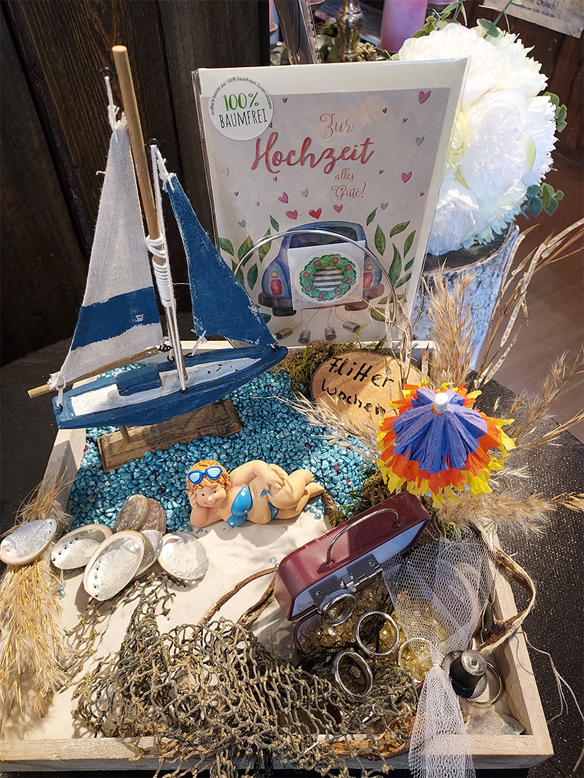 Ein maritimes Dekor mit Segelboot, Muscheln und Figur auf blauem Kies.