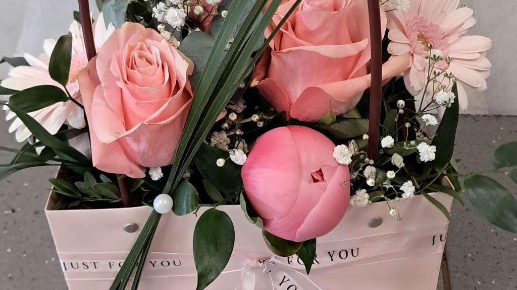 Ein Blumenarrangement mit rosa Rosen, Gerbera und zartem Grün in einer rosa Geschenktasche.