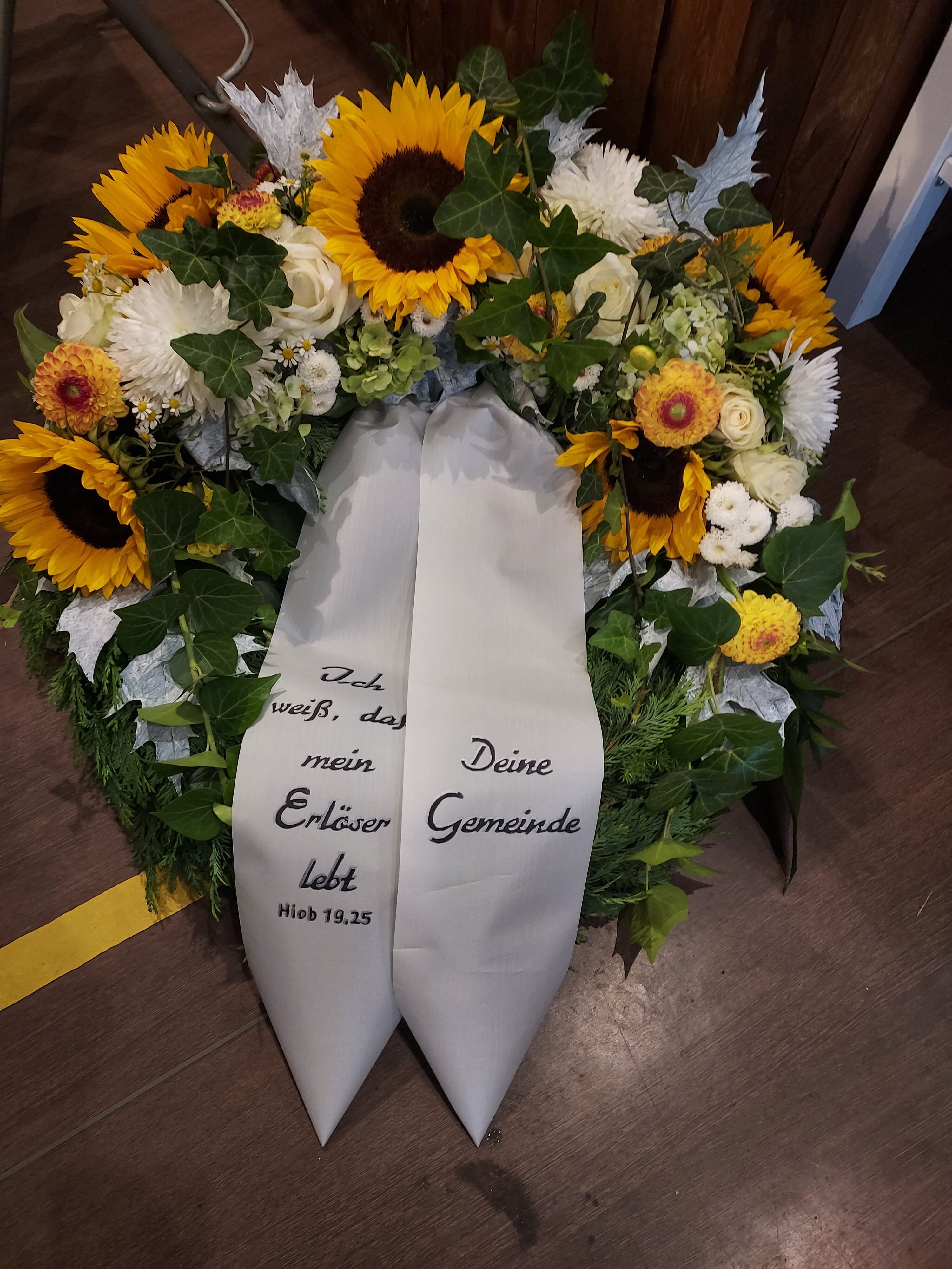 Ein bunter Blumenkranz mit Sonnenblumen und einem weißen Trauerband in einer Kirche.