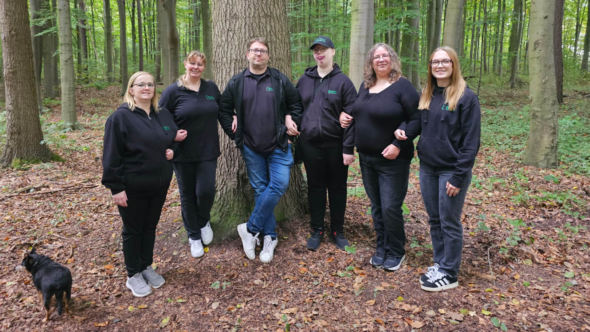 Sechs lächelnde Personen in schwarzen Hoodies stehen im grünen Wald um einen Baum